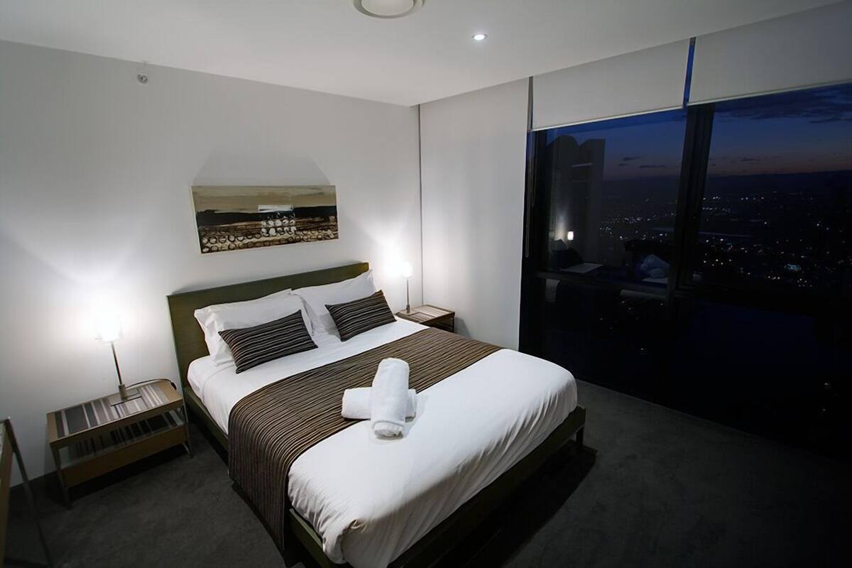 A modern hotel room with a large bed, a nightstand, a lamp, and a city view through large windows.