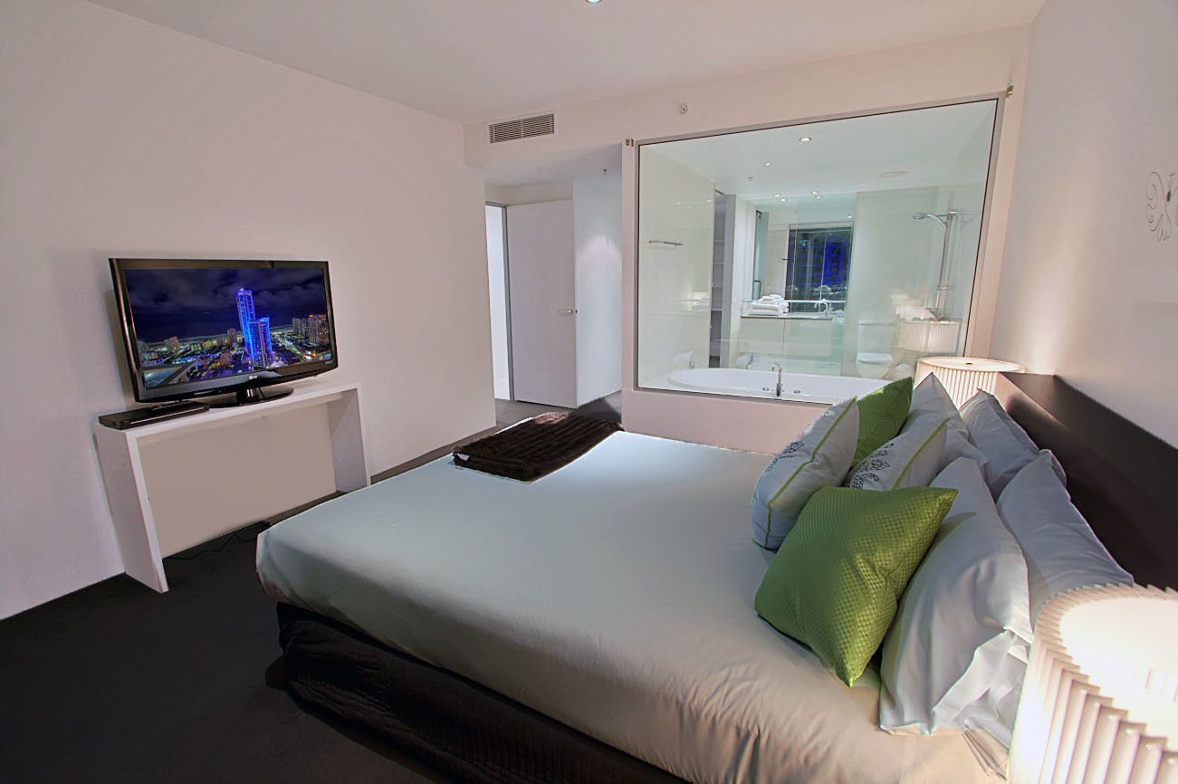 A hotel room with a bed, a TV on a stand, a large mirror, and a view of a bathroom.