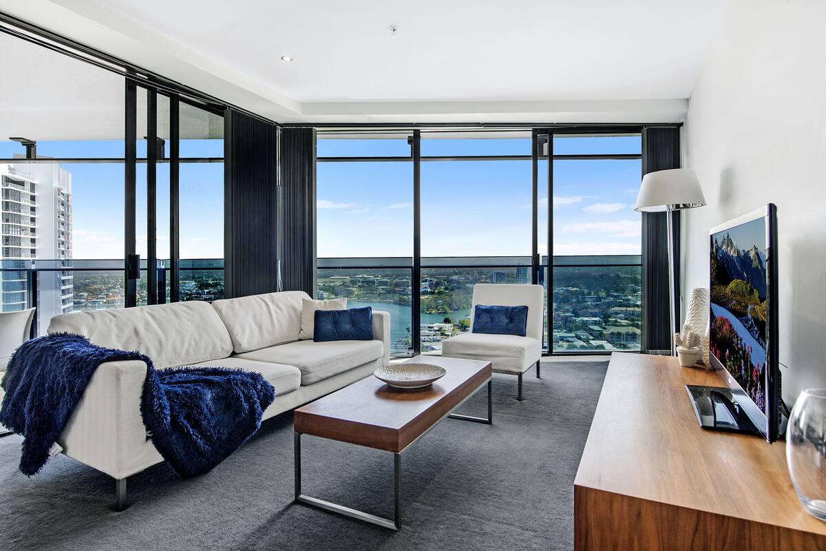 A modern hotel room with a large window, a sofa, a coffee table, and a desk with a lamp.
