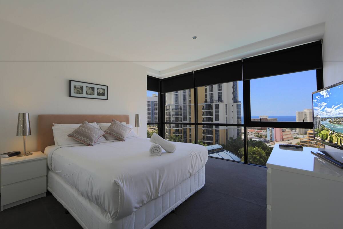 A modern hotel room with a large bed, a nightstand, a desk, and a view of the cityscape through large windows.