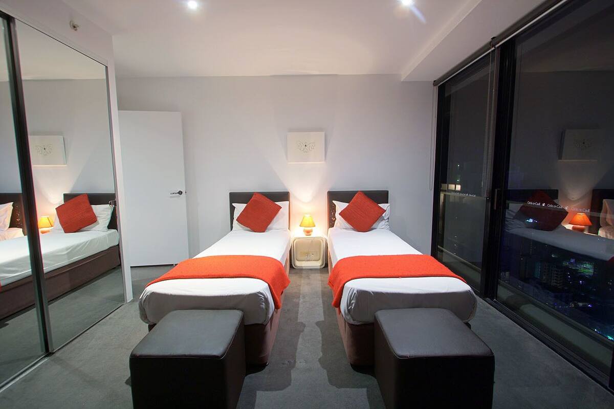 A modern hotel room with two beds, a large mirror, a city view, and a sliding door.
