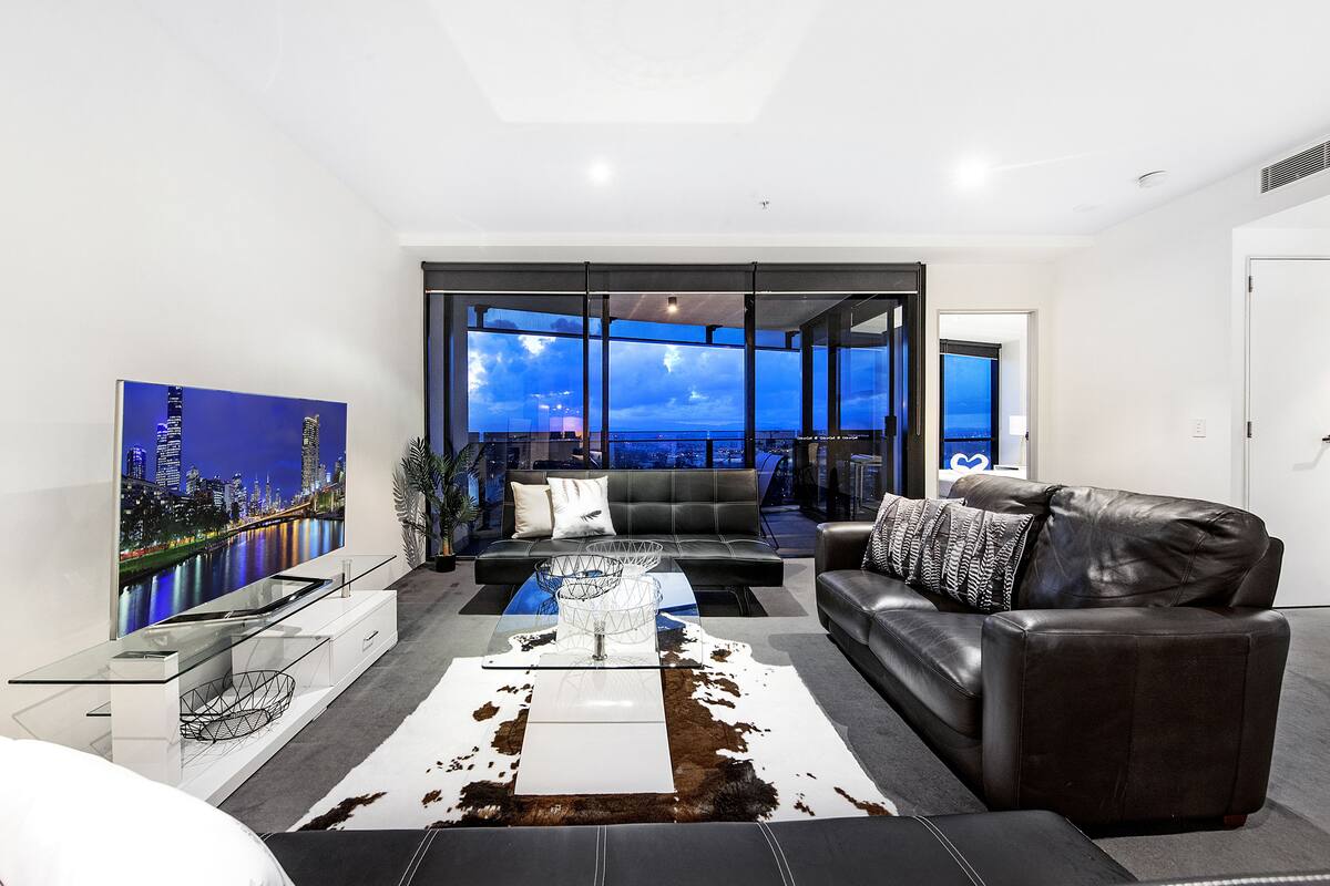 A modern living room with a large flat-screen TV, leather sofas, a glass coffee table, and a cityscape view through large windows.