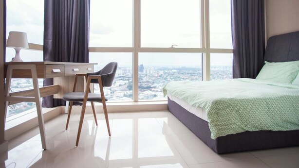 A bedroom with a bed, a desk, and a chair, large windows with curtains, and a view of a cityscape. - Pinnacle Studio - Heart of Petaling Jaya (Petaling Jaya)