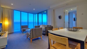 A hotel room with a large window offering a view of the ocean, a dining area with a table and chairs, and a seating area with striped chairs. - Orchid - Private Apartments (Surfers Paradise)