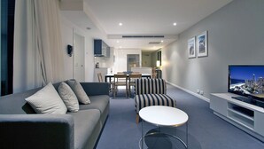 A modern living room with a sofa, armchairs, a coffee table, and a TV. There is a dining area with a table and chairs in the background. - Orchid - Private Apartments (Surfers Paradise)