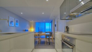 A modern kitchen with a dining area, featuring a large window with a city view, white cabinetry, and a stainless steel range hood. - Orchid - Private Apartments (Surfers Paradise)