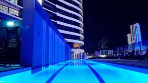 Una piscina con iluminación azul, un edificio moderno con balcones y un cielo nocturno despejado.