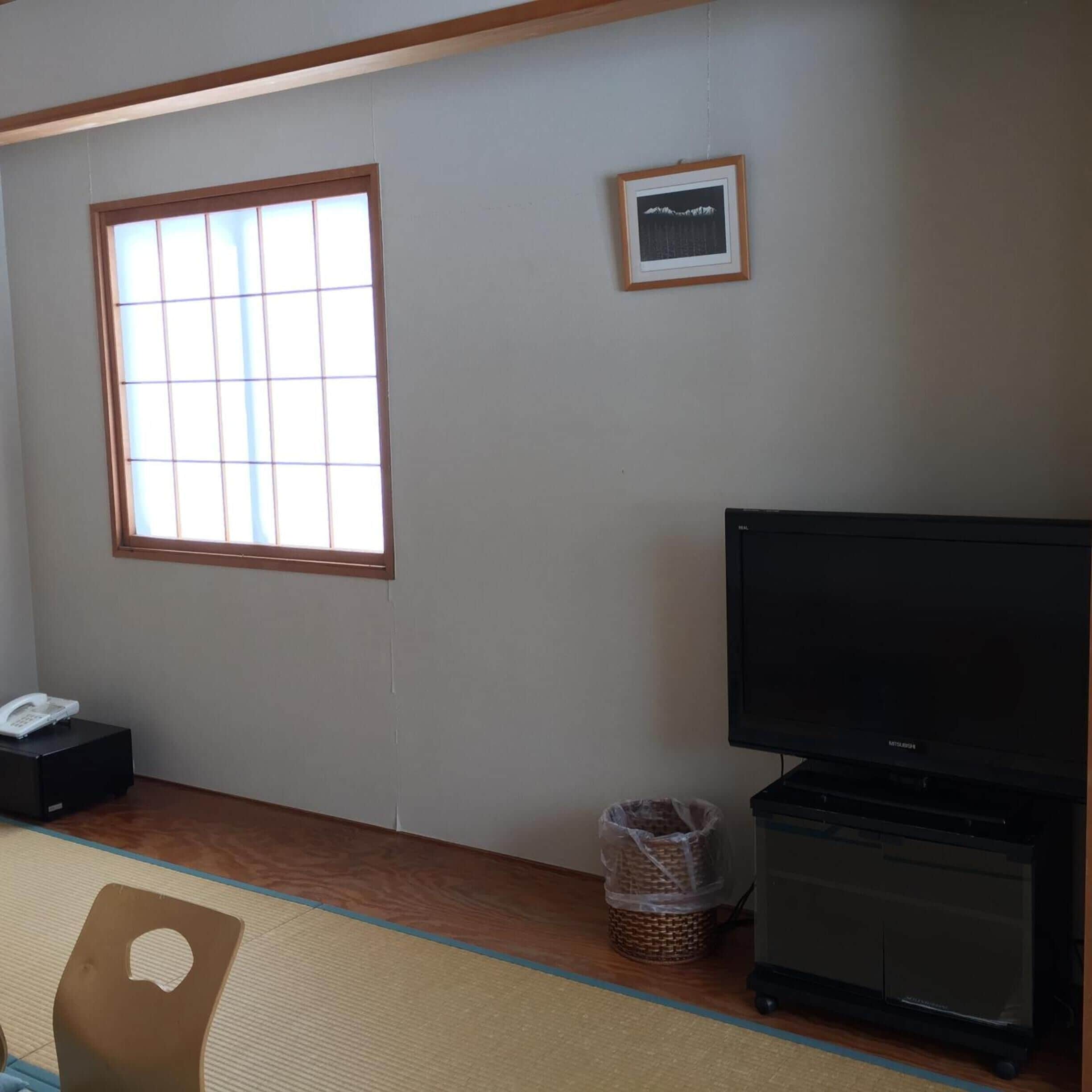 A room with a large window, a framed picture on the wall, a television, a phone, and a small basket.