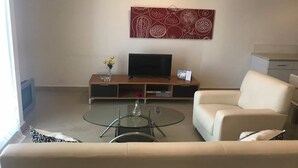 Ein modernes Wohnzimmer mit einer Couch, einem Glastisch, einem Flachbildfernseher auf einem Holzständer und einer an der Wand montierten Uhr.