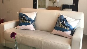Ein beiges Sofa mit gemusterten Kissen, ein Glastisch und eine Vase mit einer violetten Blume.