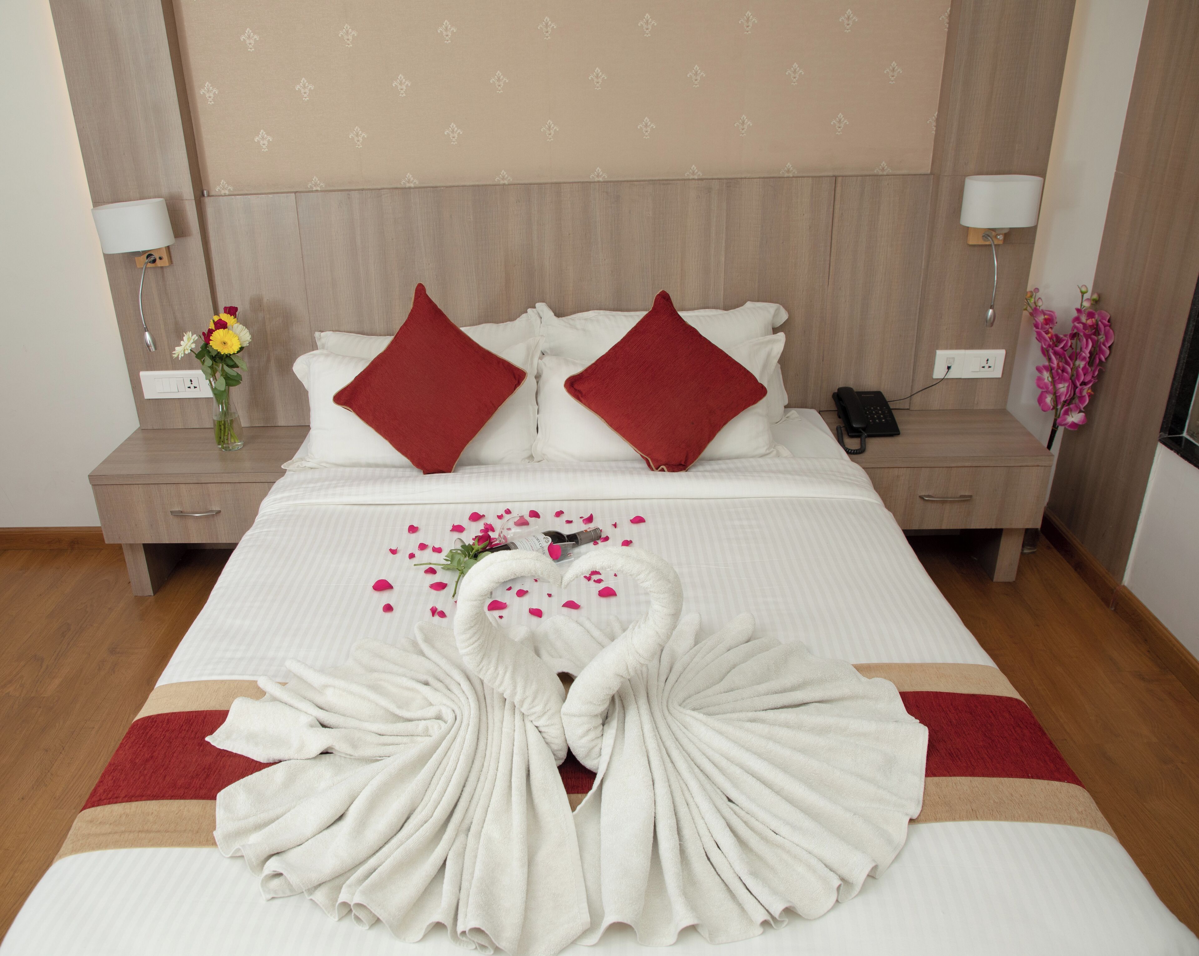 A neatly made bed with red pillows, a vase of flowers, and a swan towel arrangement.