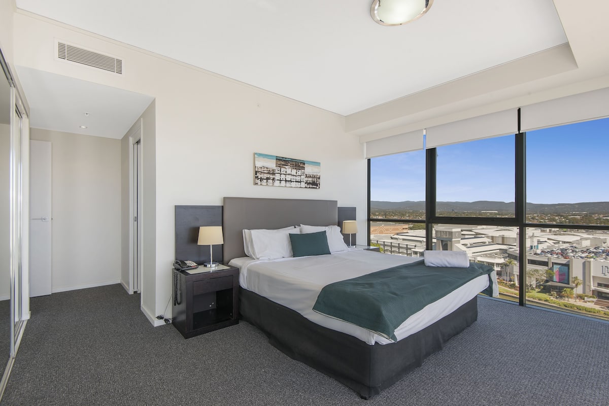 A modern hotel room with a large bed, bedside lamps, a telephone, and a city view through large windows.