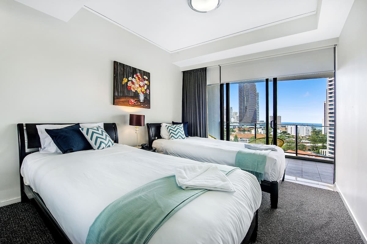 A hotel room with two beds, a painting on the wall, a nightstand, a lamp, and a view of the city through large windows.