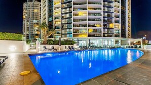 A nighttime view of a hotel with a large indoor swimming pool surrounded by lounge chairs and a modern building in the background.