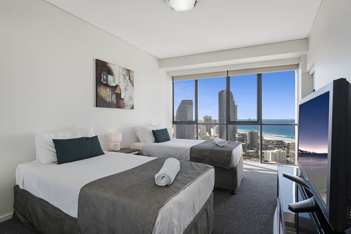 A hotel room with two beds, a large window with a city and beach view, and a television.