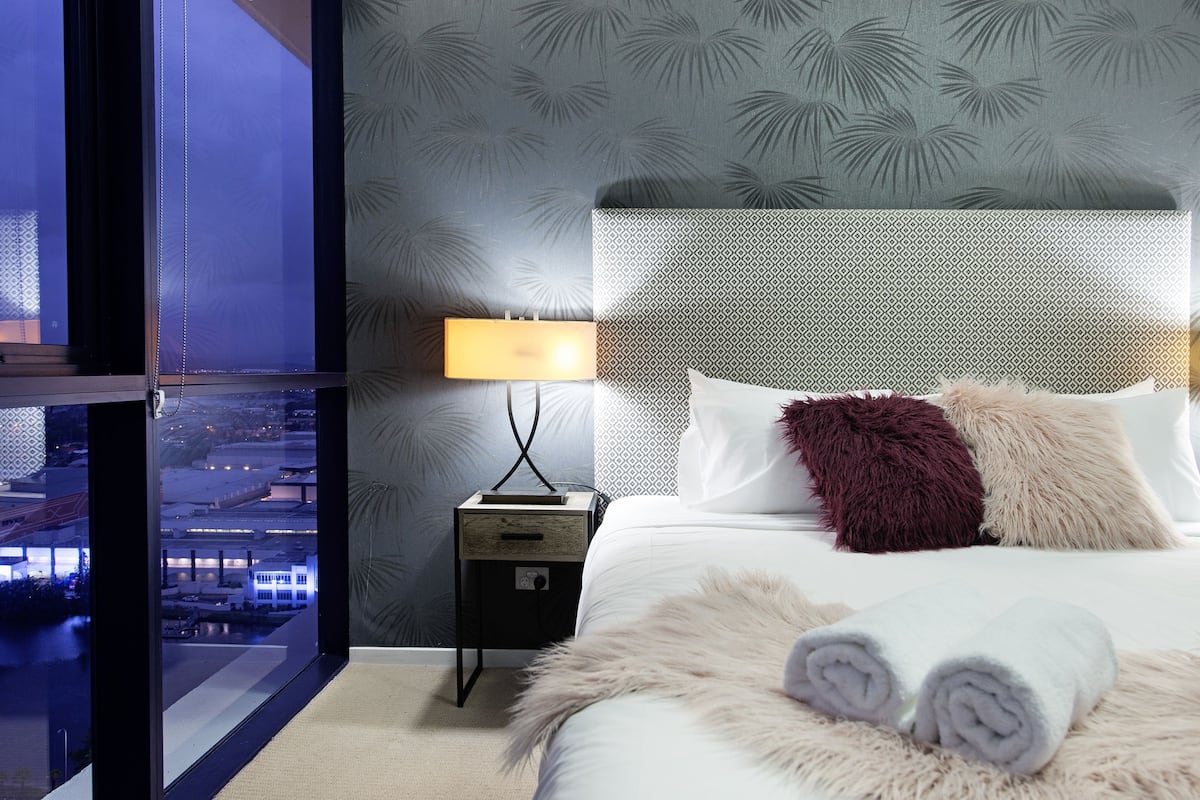 A modern hotel room with a large window offering a city view, a neatly made bed with fluffy pillows, and a small bedside table with a lamp.