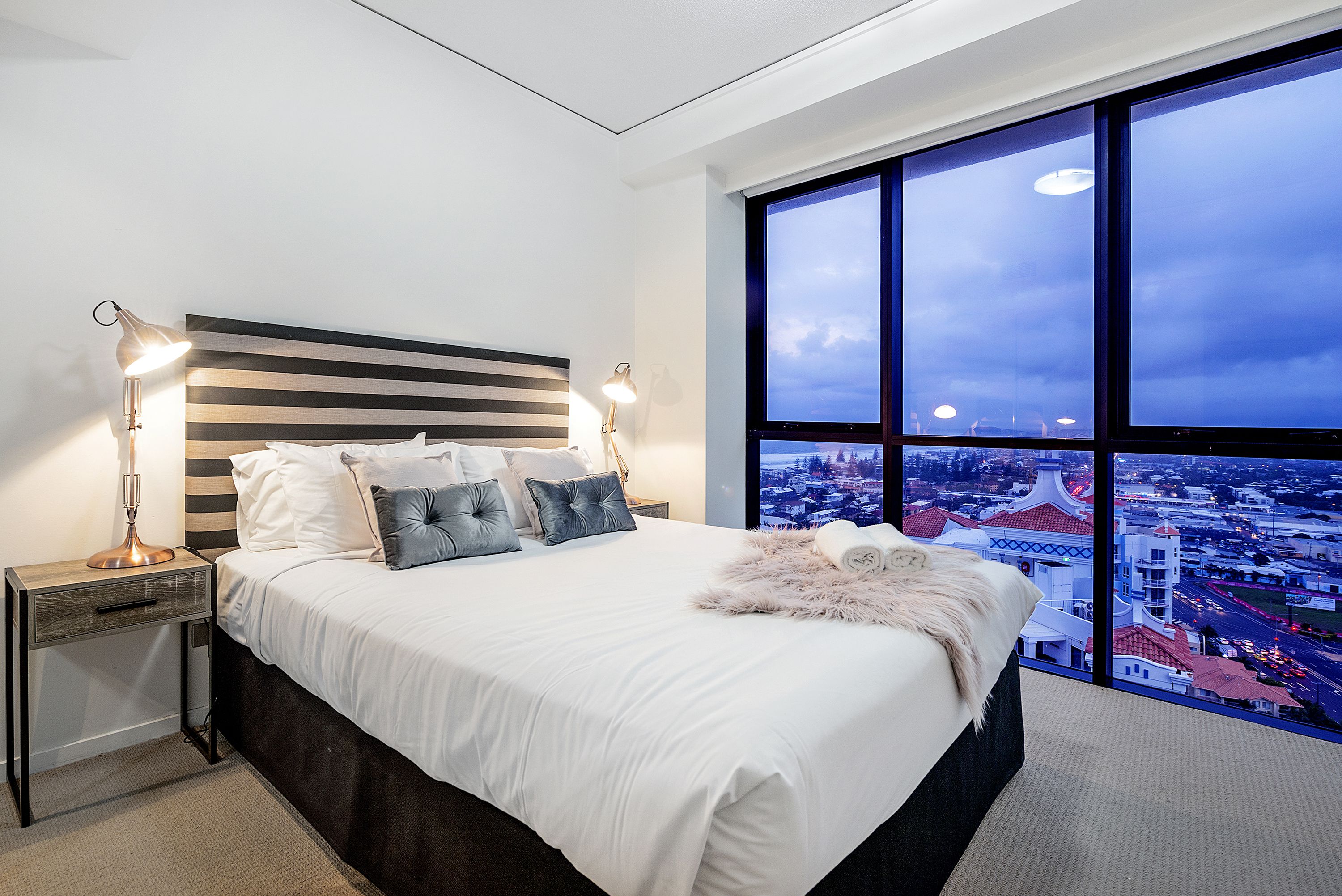 Une chambre moderne avec un grand lit, deux tables de chevet avec des lampes, une vue sur la ville à travers de grandes fenêtres et une vue panoramique sur le paysage urbain depuis la fenêtre.