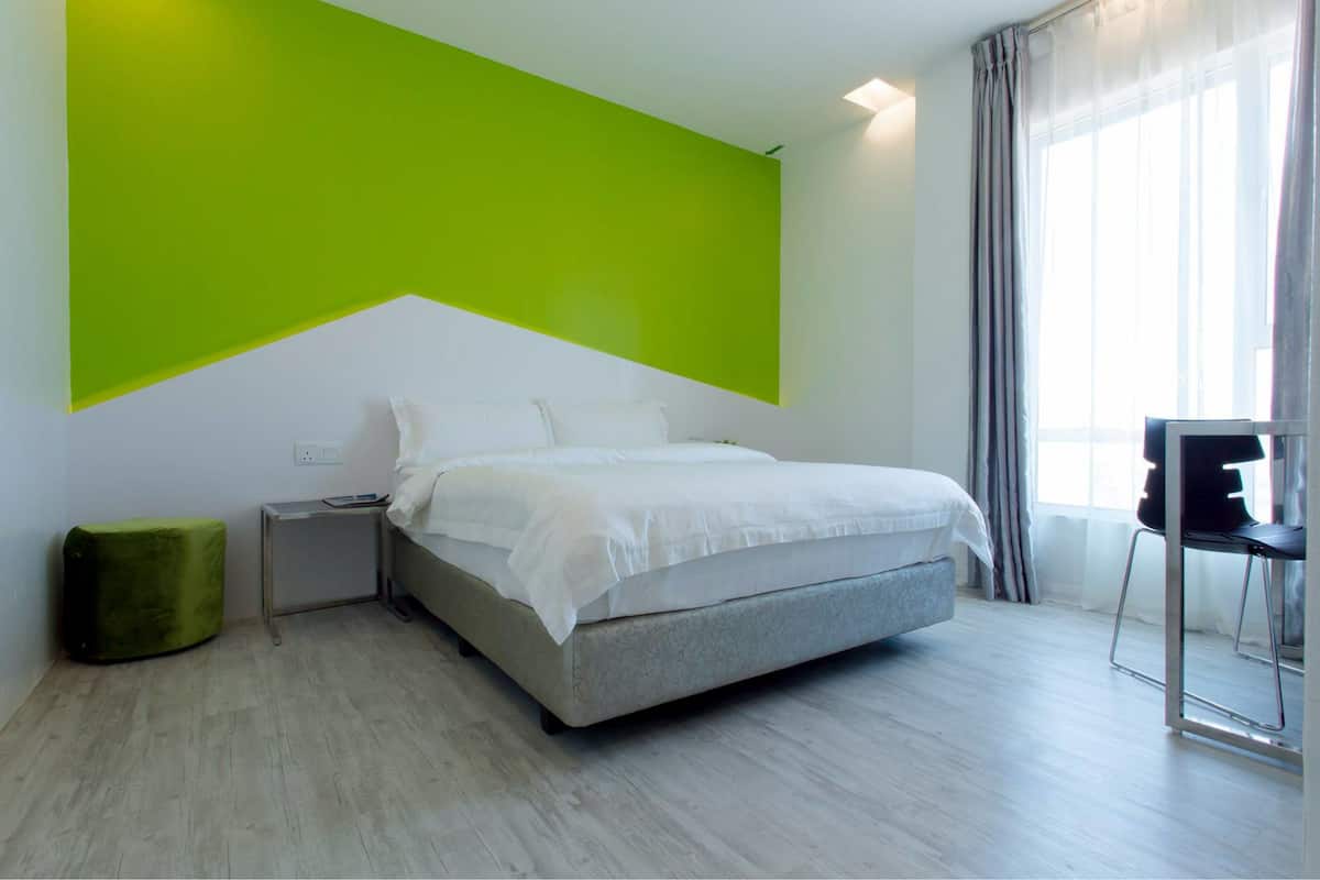 A modern bedroom with a bed, a small bedside table, a green ottoman, and a chair.
