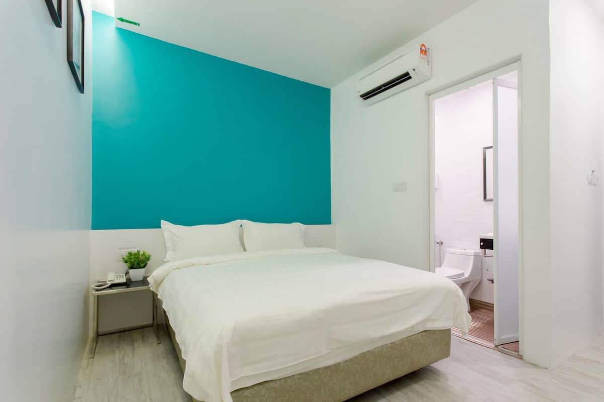 A bedroom with a bed, a nightstand, a potted plant, and a bathroom visible through an open door.