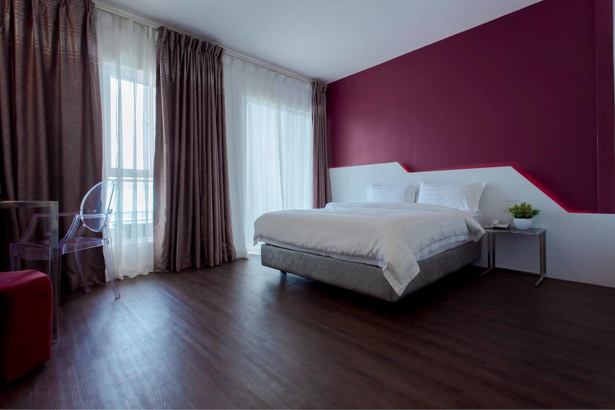 A modern bedroom with a large bed, a nightstand, a red accent wall, and a view of the outdoors through sheer curtains.
