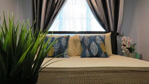Una cama con almohadas estampadas, una planta en maceta y una ventana con cortinas.