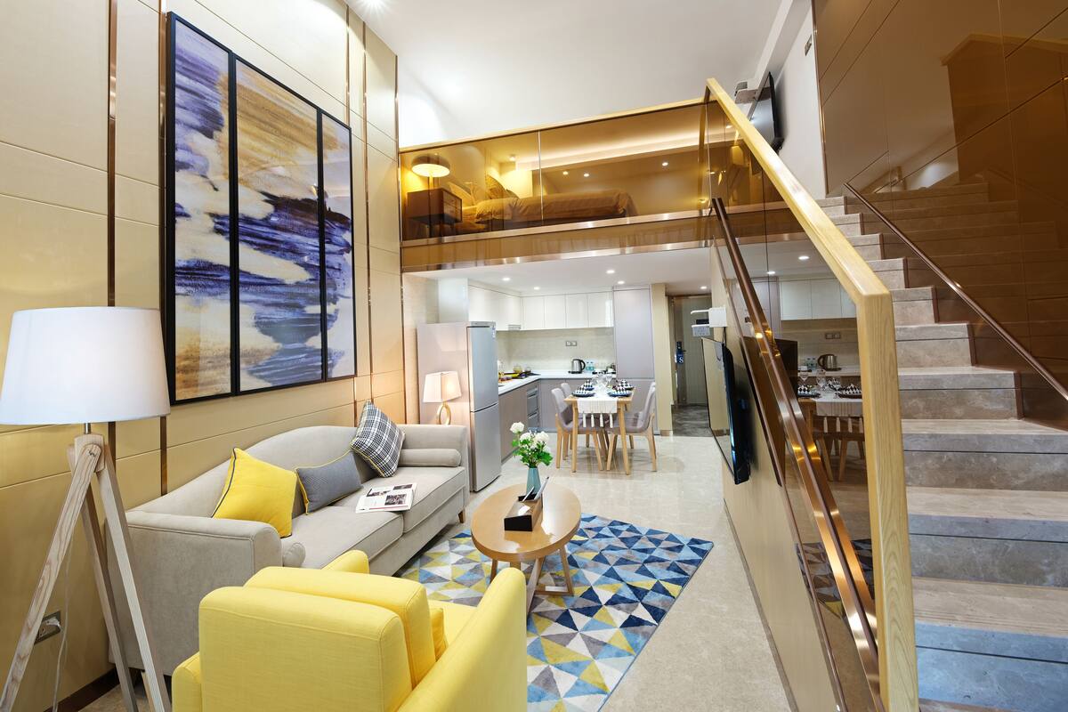 A modern living room with a sofa, yellow chairs, a coffee table, and a painting on the wall. A staircase leads to an upper level.
