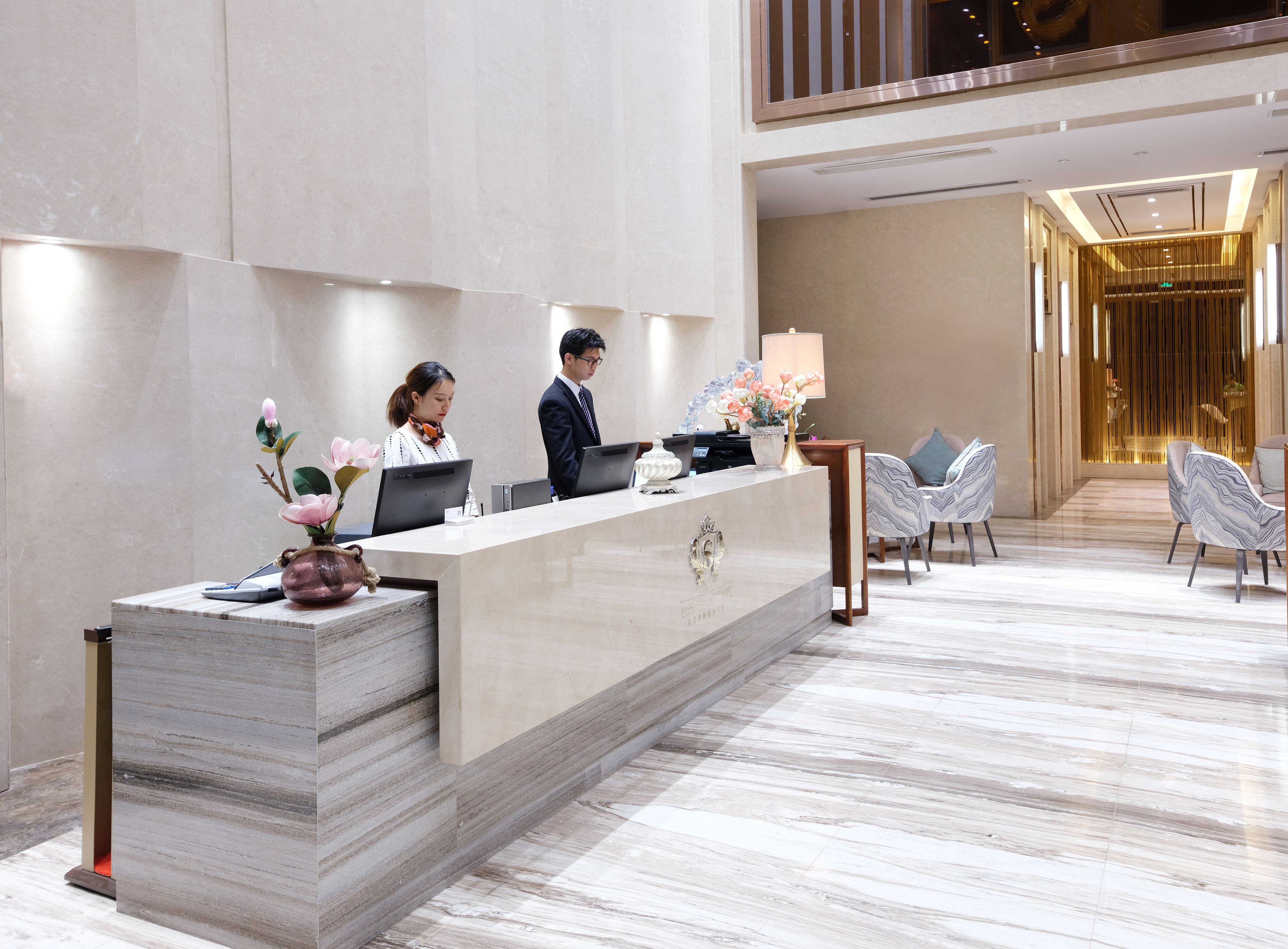 A hotel reception area with a marble reception desk, two staff members, and modern seating.