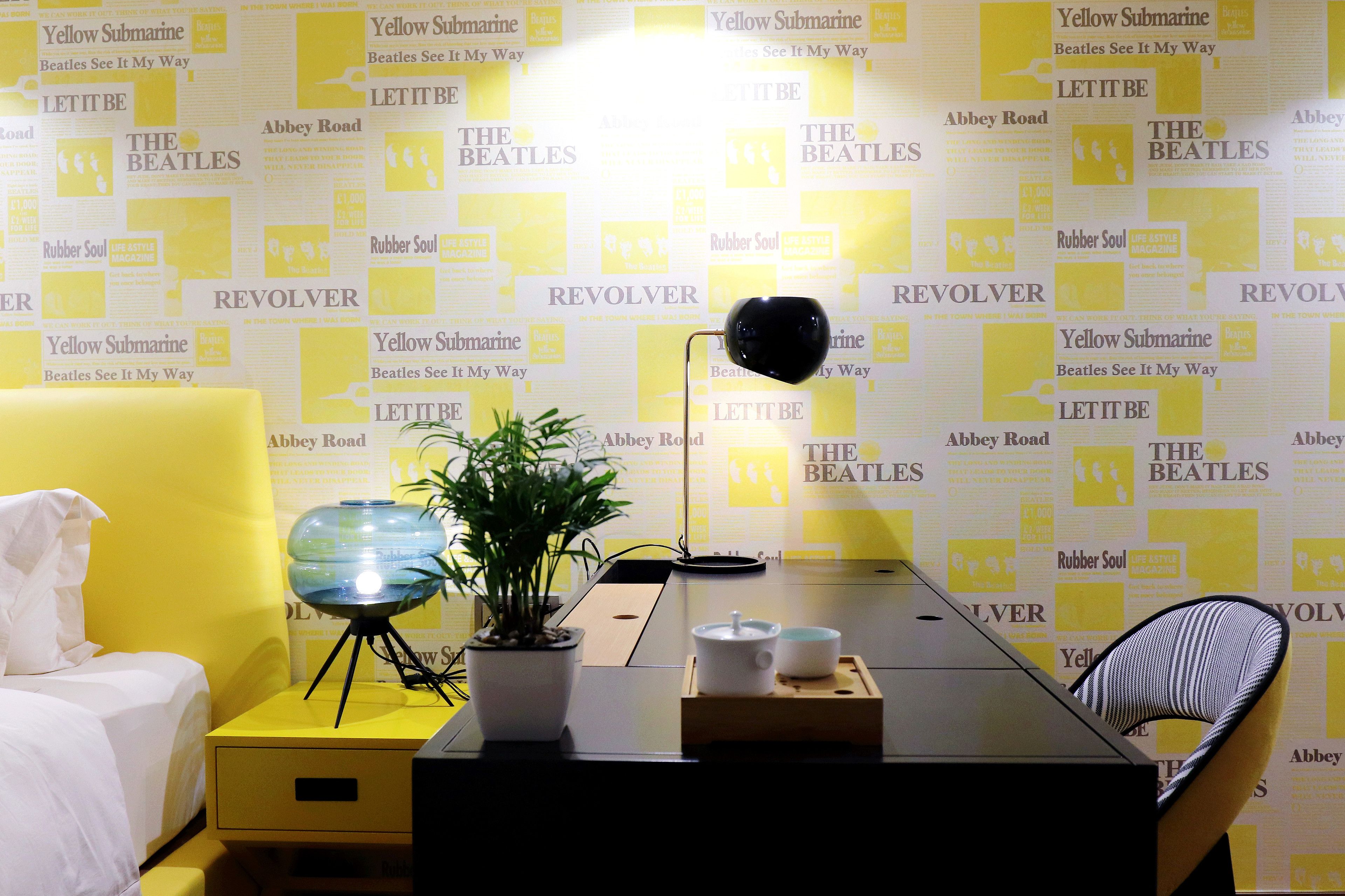 Habitación de hotel con pared amarilla, cama, escritorio con lámpara, planta en maceta y una silla.
