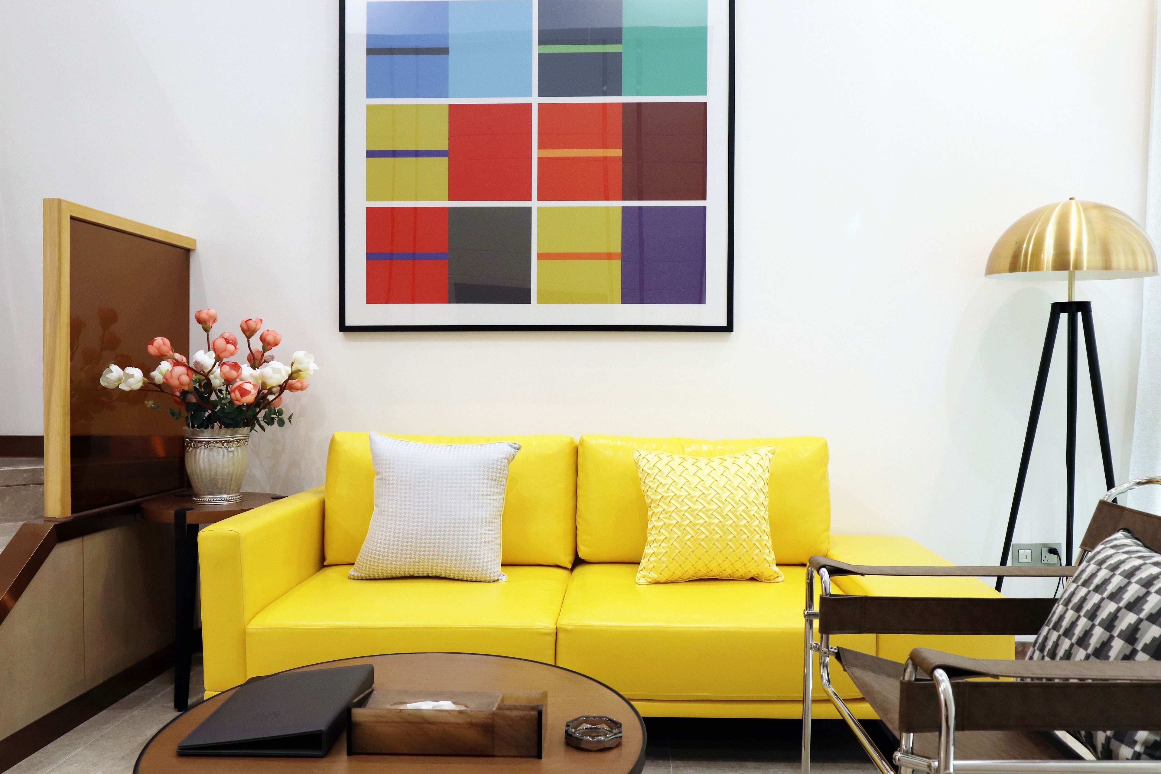 A yellow sofa with patterned cushions, a framed abstract painting, a vase with flowers, and a floor lamp.