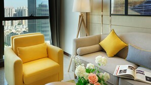 Een moderne woonkamer met een gele fauteuil, een beige bank met gele en blauwe kussens, een vloerlamp en een vaas met bloemen.
