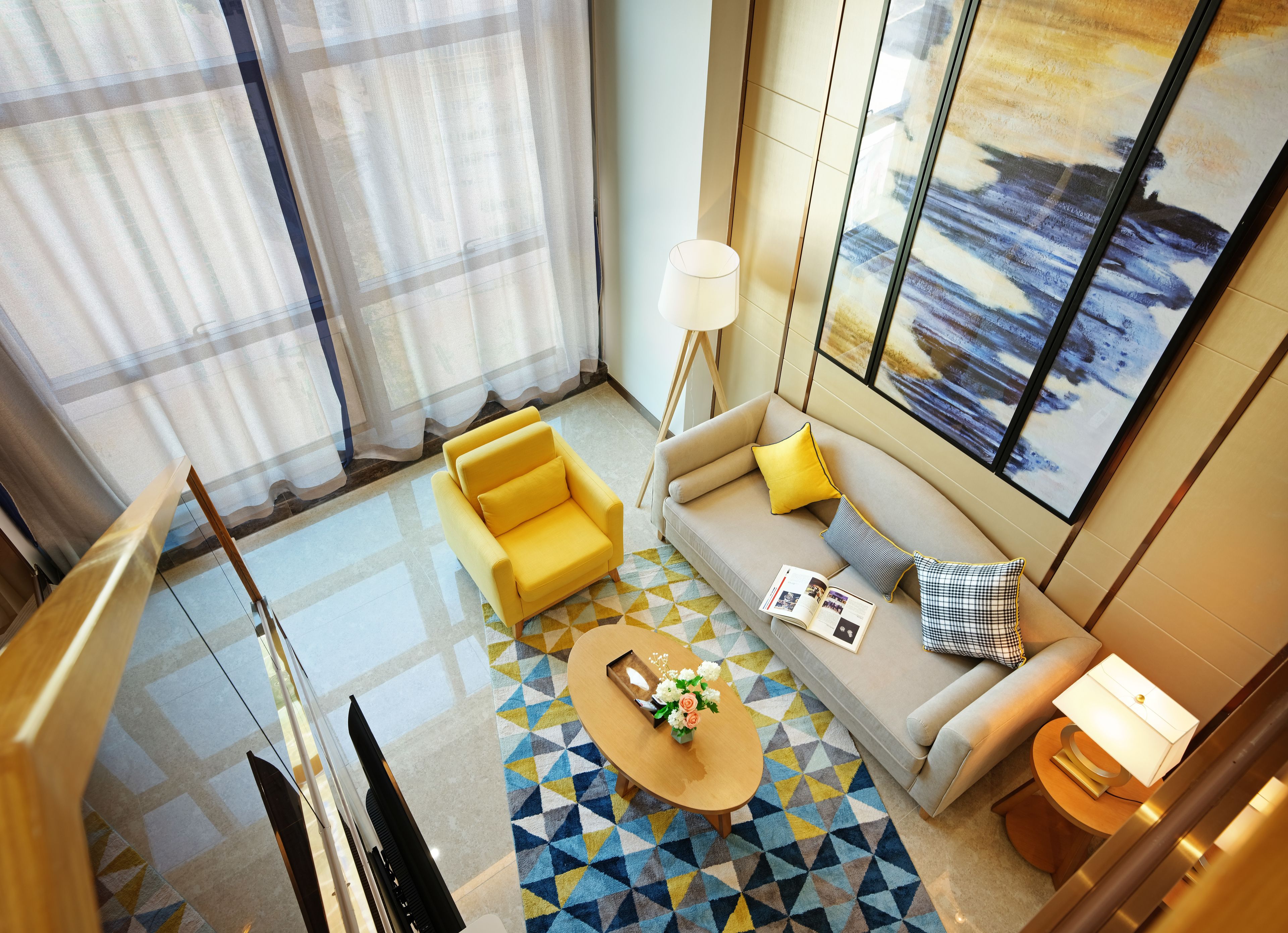 A modern hotel room with a sofa, a coffee table with flowers, a patterned rug, and a large window with a painting.