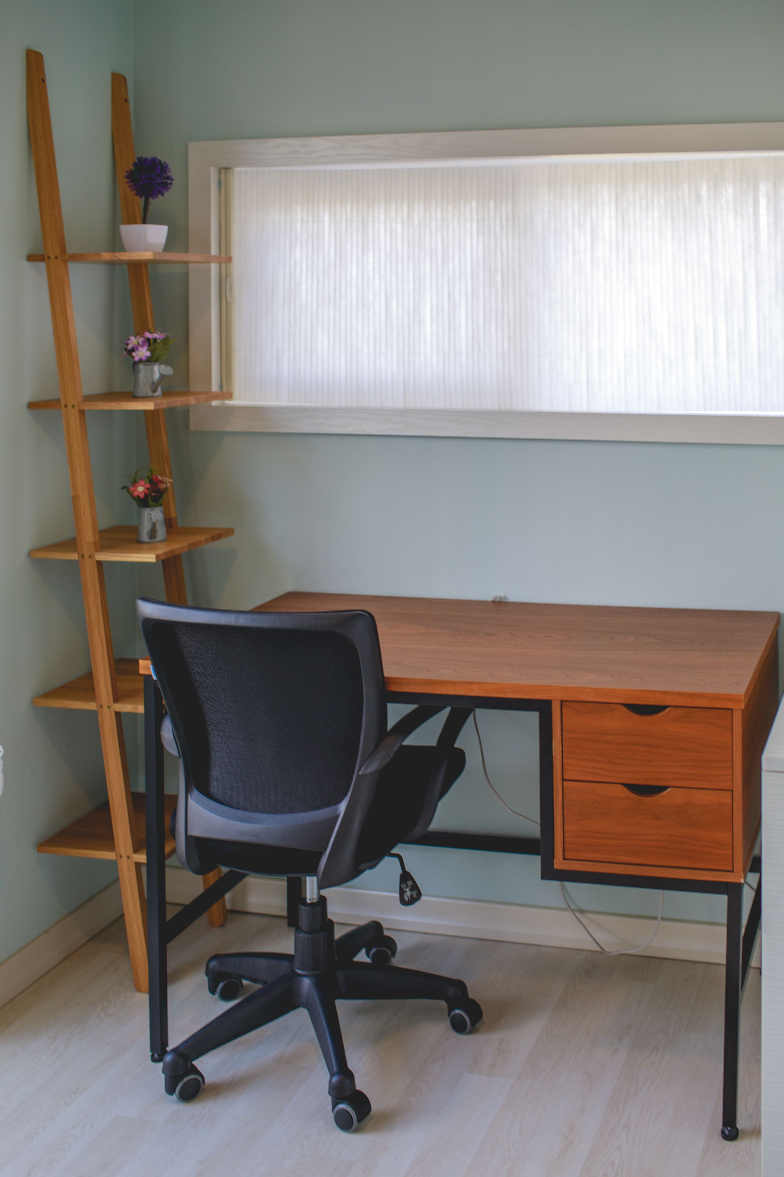 A wooden desk with a black office chair, a wooden ladder, and a window with blinds.