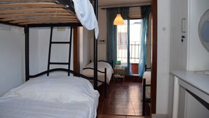 A dormitory room with bunk beds, a small table with a lamp, and a view of the outside through a window with curtains.