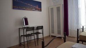 A room with a bed, a desk with two chairs, a wardrobe, a painting of a bridge, and a window with curtains.