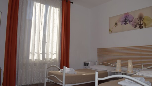 A hotel room with two single beds, a large window with orange curtains, a wall-mounted air conditioner, and a painting of flowers.