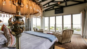 Quarto com uma cama grande, um candelabro e vista para o exterior através de janelas amplas.