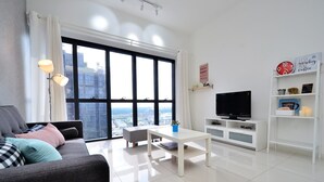 Un salon moderne avec un canapé gris, une table basse blanche, un téléviseur à écran plat posé sur un meuble blanc et une grande fenêtre donnant sur la ville.