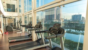 Un gimnasio con cintas de correr y equipamiento de ejercicio, amplias ventanas con vista a la ciudad, y suelo de madera.