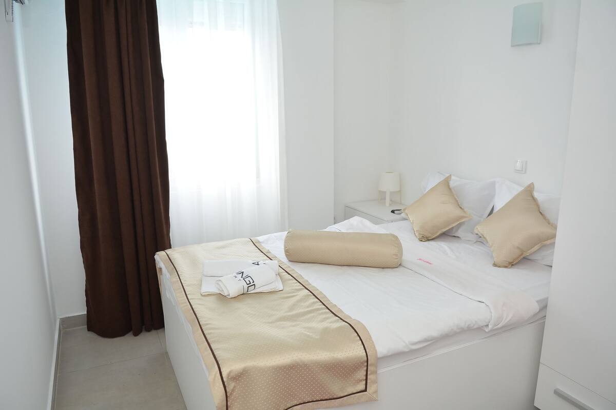 Una cama bien hecha con una manta color beige y almohadas, una mesita de noche blanca con una lámpara y una ventana con cortinas marrones.