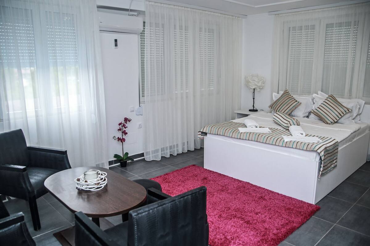 Un dormitorio moderno con cama, una mesita y sillas. Se ve una puerta que da a otra habitación.