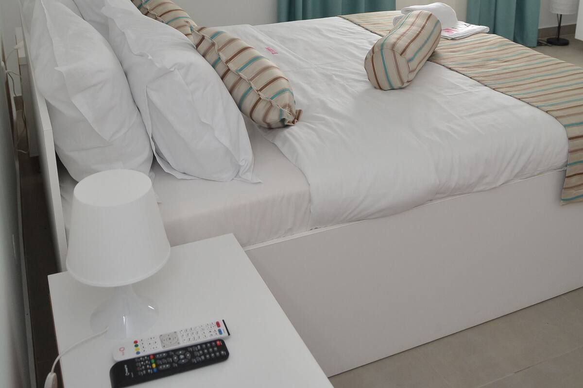 Una cama bien tendida con ropa de cama blanca y almohadas a rayas, una mesita de noche blanca con un control remoto y cortinas verde azulado en la ventana.