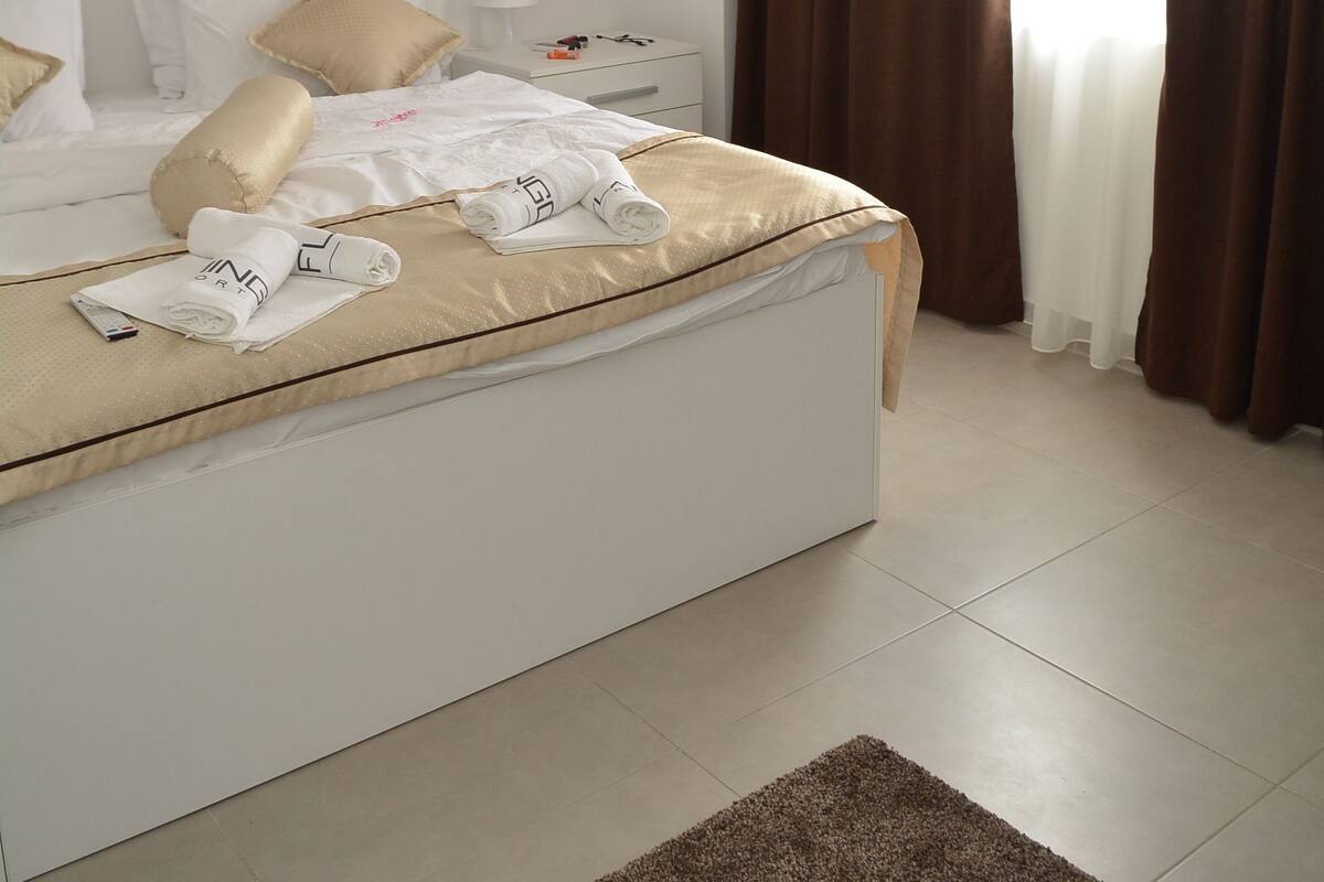 Una cama bien hecha con sábanas blancas y un plaid color beige, una mesita de noche con una lámpara y una ventana con cortinas marrones.