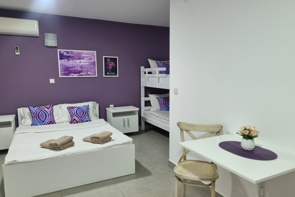 Une chambre d’hôtel avec un lit superposé, un lit simple, un bureau avec une chaise et une petite table sur laquelle est posé un vase de fleurs.