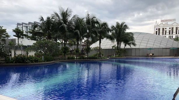 Ein Schwimmbecken mit klarem, blauem Wasser, umgeben von Palmen und einem modernen Gebäude mit Kuppelbau im Hintergrund.