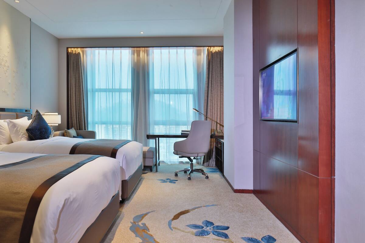 Habitación de hotel con dos camas, un escritorio, una silla, un televisor y ventanas amplias con cortinas.