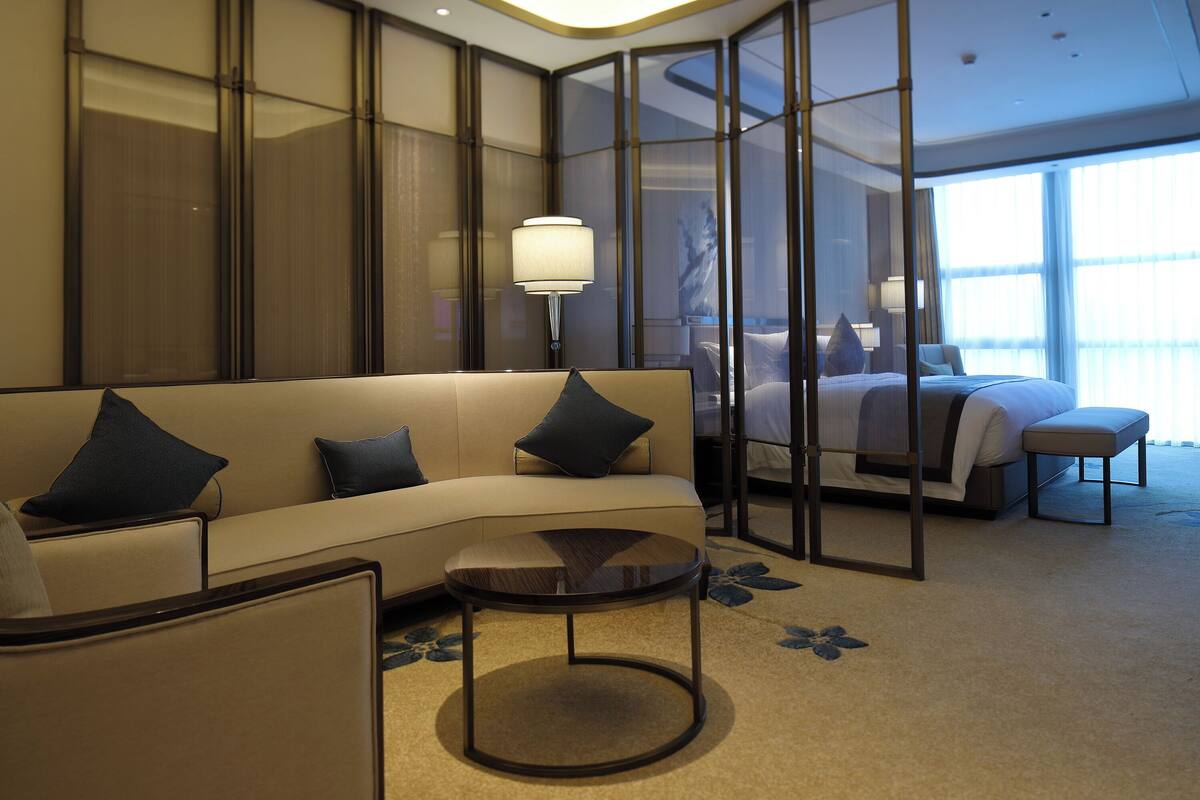 Una habitación de hotel moderna con sofá, una mesita redonda, cama y un ventanal que proporciona luz natural.