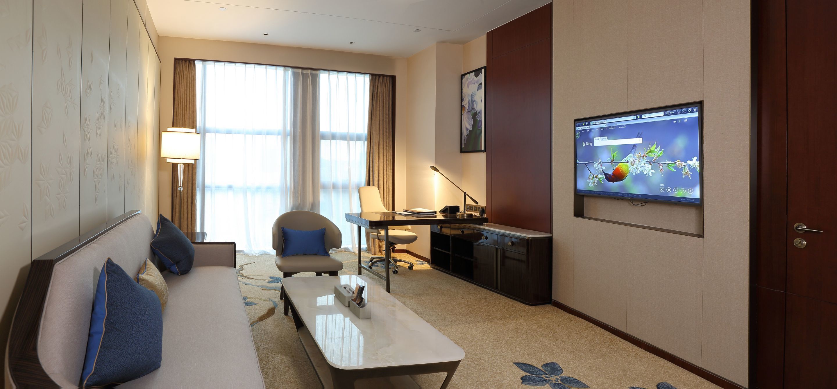 A hotel room with a sofa, a desk, a chair, a TV, and a large window with curtains.