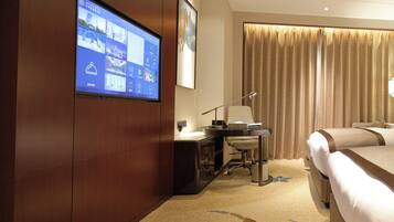 A hotel room with a bed, a desk, a chair, a TV mounted on the wall, and a large window with curtains.