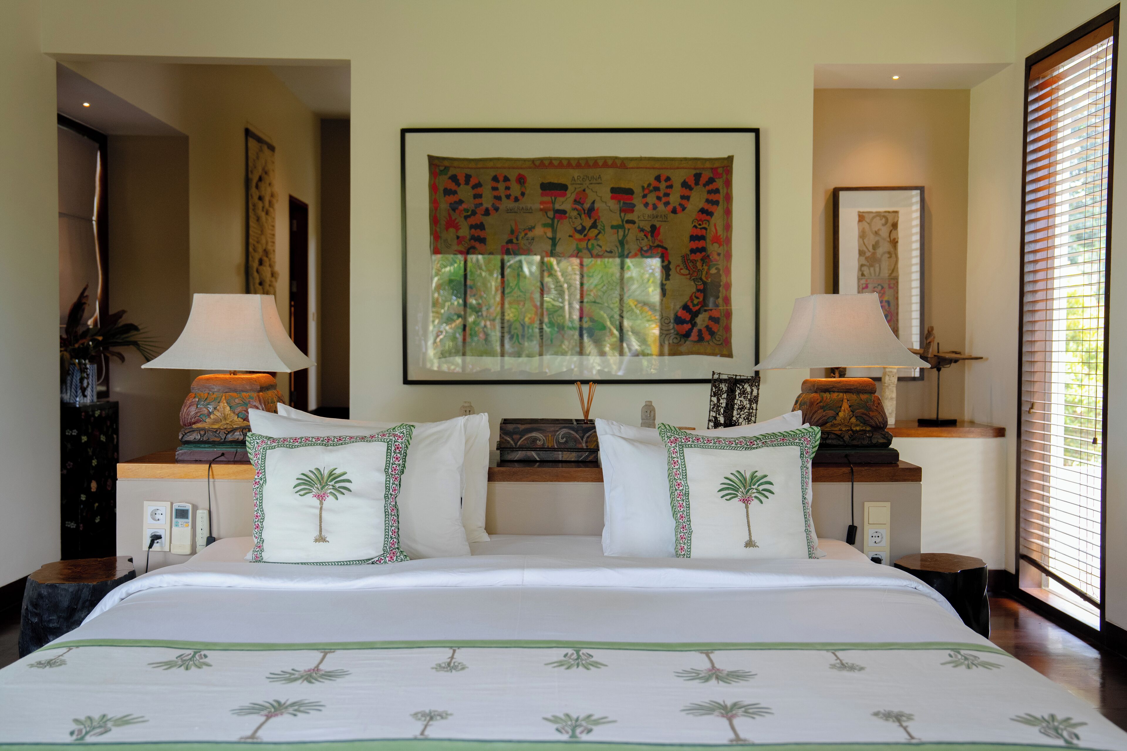 A hotel room with a large bed, two bedside tables with lamps, a wall tapestry, and a window with blinds.