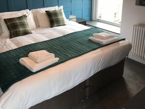 A neatly made bed with white linens and a green throw blanket, a bedside table, and a window with a view of buildings.